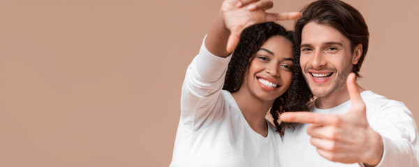 Fototapeta premium Capturing Happy Moment. Romantic Multiracial Couple Framing Their Faces With Fingers nd Sincerely Laughing, Posing Together On Yellow Background