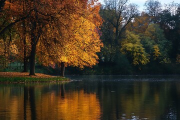 Fototapeta premium Autumn tree reflecting in still lake water with golden leaves and green woods in background.
