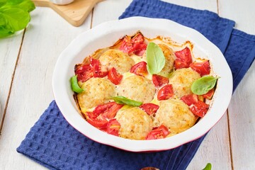 Fish meatballs baked with tomatoes and sauce in a ceramic baking dish on a white wooden background.