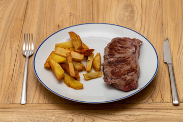 a white plate with a blue ring with a grilled beef steak and French fries on a wood board