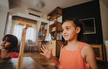 Two young girls play enthusiastically with wooden blocks in a cozy living room setting. Their...