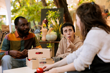 Caucasian farmer offering freshly harvested bio produce to multiracial customers. Young diverse consumers seated and tasting locally grown organic fruits at harvest farm fair festival.
