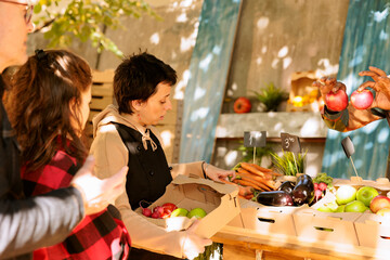 Female merchant selling natural fruits and veggies in boxes, serving senior man and young woman at farmers market. Young vendor picking various freshly harvested produce for customers.