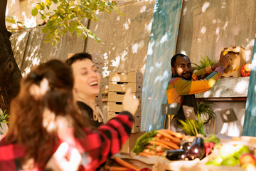 African American farmers market vendor holding lemons and smiling to customers, standing behind the fruits and vegetables stand. People are purchasing fresh bio organic produce grown locally.