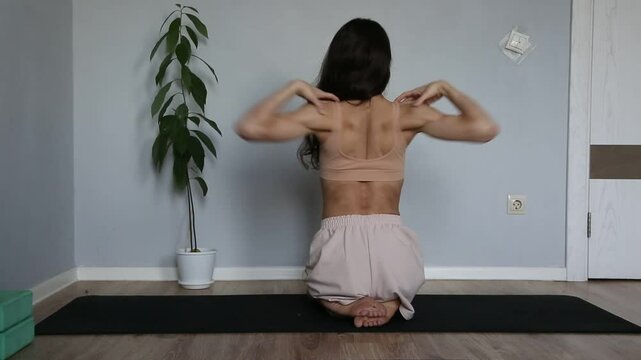 Beautiful fit atletic body woman doing stratching exersices for shoulders, scapula and healthy back in bra and sporty wear sitting on black mat at home interior. Health yoga and sport lifestyle