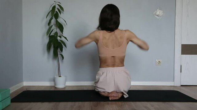 Beautiful fit atletic body woman doing stratching exersices for shoulders, scapula and healthy back in bra and sporty wear sitting on black mat at home interior. Health yoga and sport lifestyle