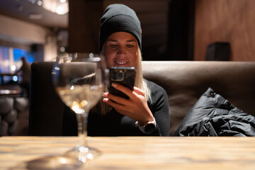 A woman in a hat in a restaurant holds a mobile phone in her hand and smiles, there is a glass of light wine on the table. Subdued lighting inside.