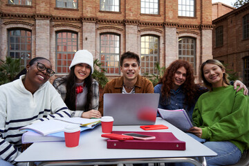 Multiethnic group of student friends gathering at university campus to study all together for a school project, sitting outside in a table using a laptop, looking at camera smiling.