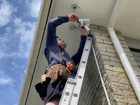 Caucasian man wearing a leather tool belt standing on a ladder installing a security sensor light to the underside of a soffit on the corner of a brick clad house in New Zealand