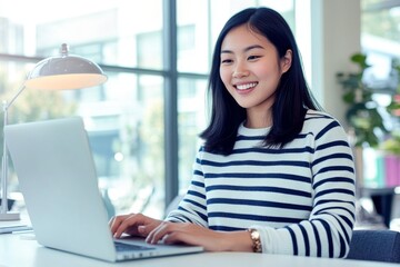 Attractive smiling Filipino woman wearing glasses and a striped sweater, sitting at her desk and typing on a computer, with a scenic window view in the background, conveying a positive and productive 