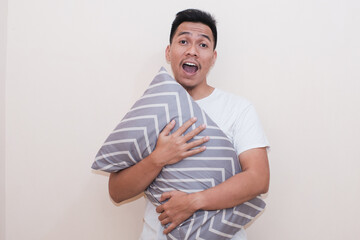 A Southeast Asian man holding pillow with expression is one of confusion or surprise, isolated over white color background, he seems to be posing in a relaxed.