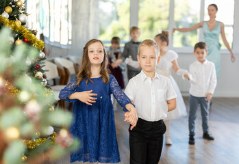 Smiling little children in elegant dresses standing in pairs in dance pose in school-hall decorated...