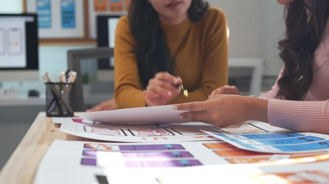 Two ux designers engage in a discussion about mobile application interface sketches laid out on a desk, collaborating on ideas and refining their creative concepts