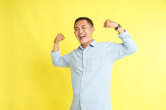 Asian man in light blue denim shirt with strong gesture and raising arm showing biceps isolated on yellow background