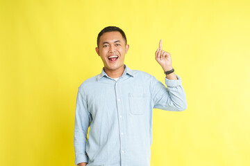 Asian man in light blue denim shirt with get idea gesture finger pointing up isolated on yellow background