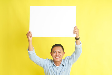 Asian man in light blue denim shirt holding blank white board for advertisement isolated on yellow background