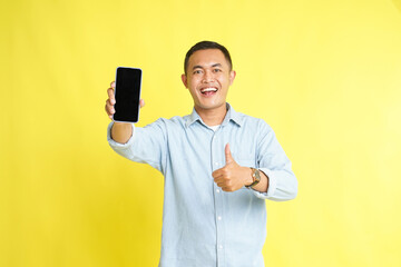 Asian man in light blue denim shirt holding blank screen phone and recommended gesture isolated on yellow background
