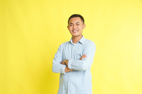 confident smiling Asian man in light blue denim shirt with arm folded isolated on yellow background
