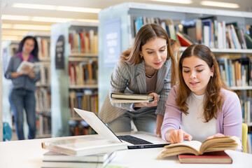 Two young women preparing to exam in library together, working on laptop computer, reading books...