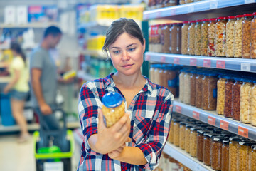 Young female shopper choosing food in supermarket, holding glass jars with canned pickled...