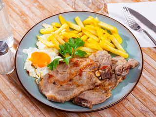 close up of grilled pork with fried potato and egg served on blue plate on wooden table.