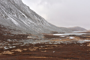 A winding river flows into a lake in a picturesque valley at the foot of a mountain range sprinkled with first snow on a cloudy autumn day.