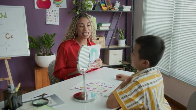 Hispanic female speech therapist conducting lesson of letters pronunciation with Asian boy in classroom decorated with educational posters