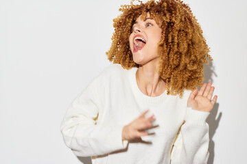 Happy woman with curly hair dancing joyfully against a plain white background, wearing a cozy white sweater, embodying a carefree and positive mood, perfect for lifestyle and happiness themes