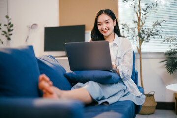 Naklejka premium Happy asian woman working from home on her laptop, sitting comfortably on a sofa, embodying a modern lifestyle