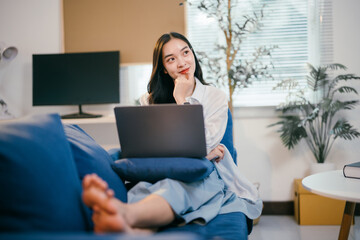 Naklejka premium Young woman sits comfortably on a cozy blue sofa in her living room, working on a laptop and lost in thought, finding inspiration in the comfort of her home