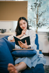 Young woman is sitting on a comfortable blue sofa in her living room, engrossed in a captivating...