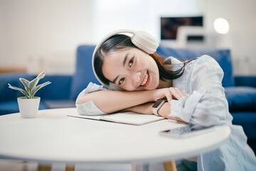 Young woman rests on a table at home, smiling contently while listening to music. She takes a break, surrounded by technology, feeling happy and relaxed in her cozy apartment