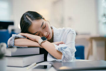 Obraz premium Exhausted young asian woman peacefully sleeps on a stack of books at her home office desk, taking a break from working on her laptop