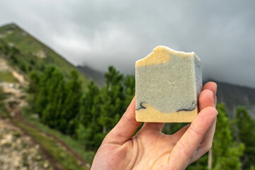 Piece of craft soap in a hand against the background of mountains, forests and sky. Concept of the image of organic cosmetics in a natural environment