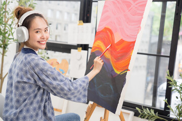 Young serious Asian woman Artist painting artwork and listen music at home workspace.