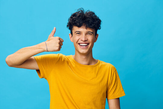 cheerful young man giving a thumbs up with a big smile, wearing a yellow t shirt on a bright blue background, radiating positivity and confidence in a casual setting