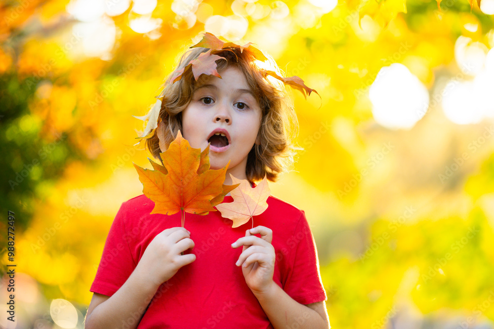 Wall mural Surprised Portrait of funny kid play autumn leaf in autumn park. Child in autumn park. Little girl with yellow leaf. Kid playing with fall leaves. Toddler kid under a maple tree on a sunny autumn day.