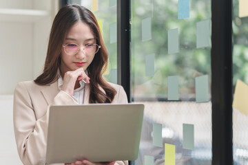 A professional woman in a suit thoughtfully engages with her laptop, surrounded by colorful sticky notes in a modern office setting.