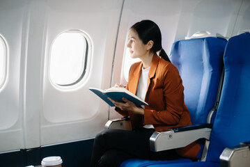 Asian businesswoman or entrepreneur in a formal suit on an airplane sitting in business class using a phone, computer laptop. Travel in style, work