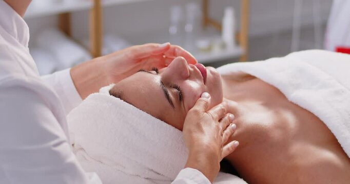 Happy young woman lying on bed at spa salon and enjoying pampering facial massage. Cosmetologist massaging face of female client for lymphatic drainage and lifting effect. Beauty, skin care concept