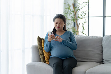 Young asian pregnant woman sitting on sofa using her mobile phone .