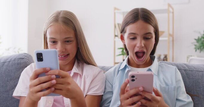 Children using smartphones. Two happy preteen girls, friends or sisters, sitting together on sofa, holding mobile phones, browsing social media, watching videos and sharing funny posts with each other