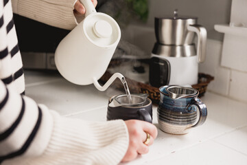 Woman in sweater pouring from a kettle into mugs with tea strainers