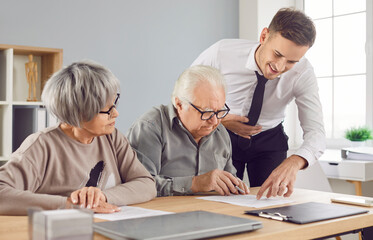 Obraz premium Confident gray-haired elderly senior couple sitting at the desk in office with a young man advisor or male financial agent signing a contract about health insurance or investments.