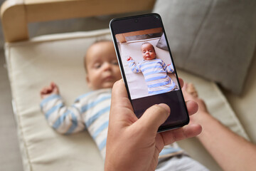 Unrecognizable anonymous man sitting on sofa with his infant son capturing cute picture for social networks while enjoying the moments of parenthood at home