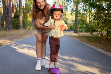 Healthy and active lifestyle in the summer. Mom and toddler spending time together outside little girl learning to ride penny board on the smooth asphalt road