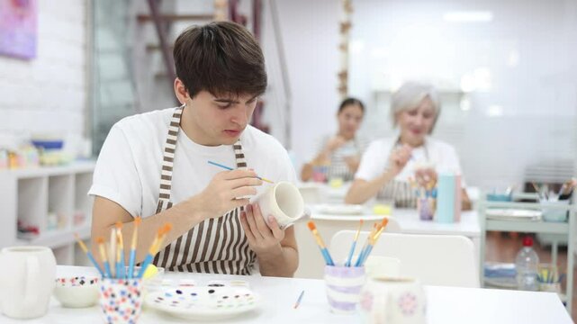 Focused young guy attending pottery class, enthusiastically painting on ceramic cup, creating unique custom design. Creative hobby concept