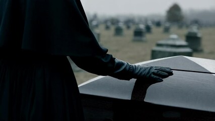Cropped shot of unrecognizable woman in long black coat putting his hand on black wooden lid of coffin at funeral outdoors