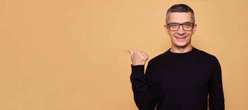 Happy Caucasian man wearing black jumper and glasses raising arm with cheerful face pointing finger to left side showing copy space for advertisement isolated over beige background