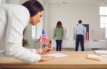 Fototapeta premium Young woman employee work in voting center. People american voters voting standing back in a row at vote center with USA flags in polling booth. Election day and democracy concept.
